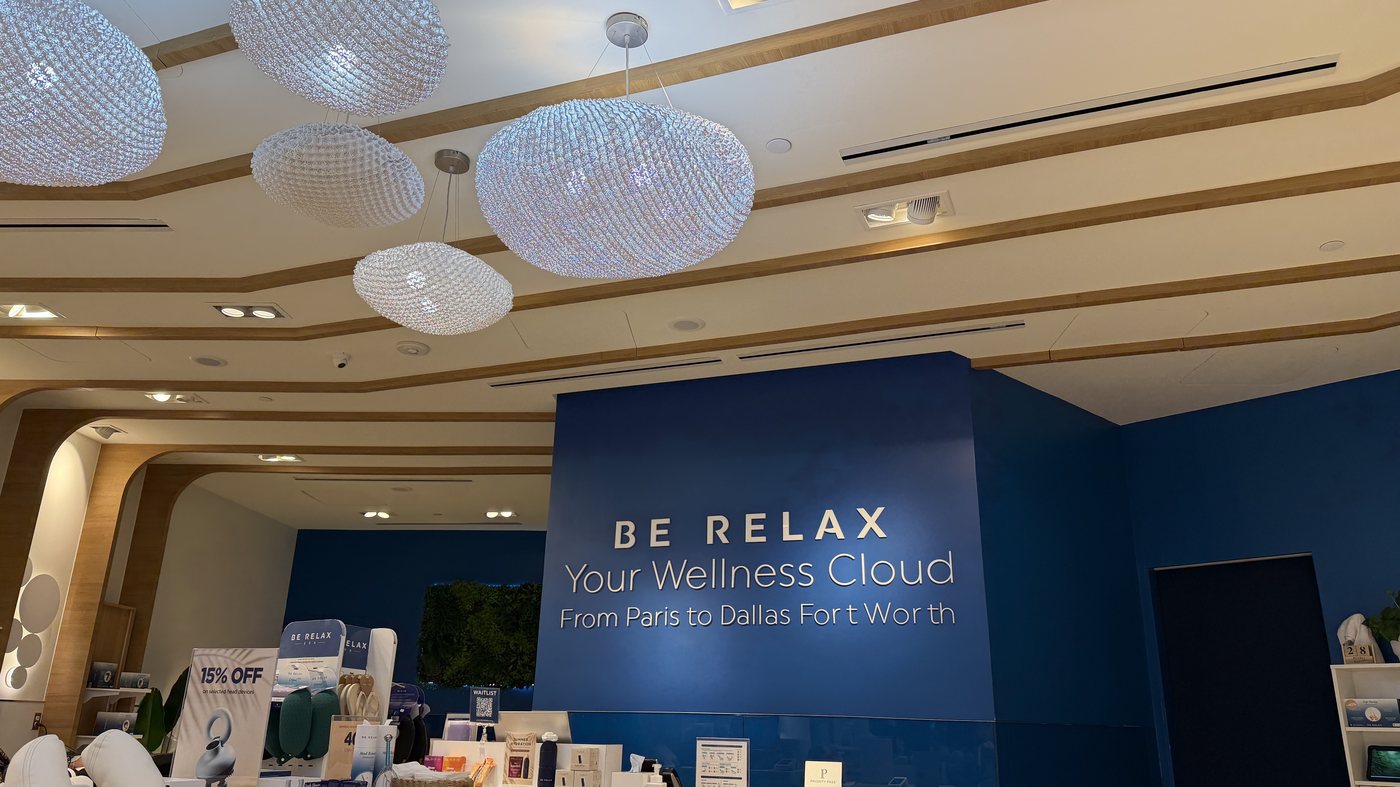 The Be Relax spa counter in DFW Terminal D, a blue wall reading "BE RELAX, Your Wellness Cloud, From Paris to Dallas Fort Worth" above a retail display
