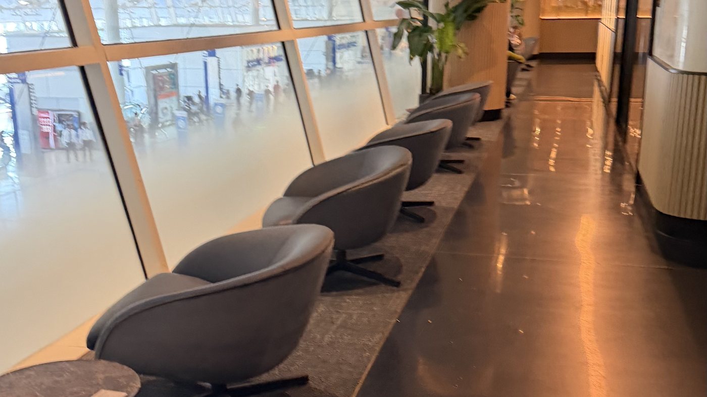 A Terminal D lounge at DFW on a weekday afternoon: a row of swivel chairs along a full-height window wall, with an empty polished walkway running past them