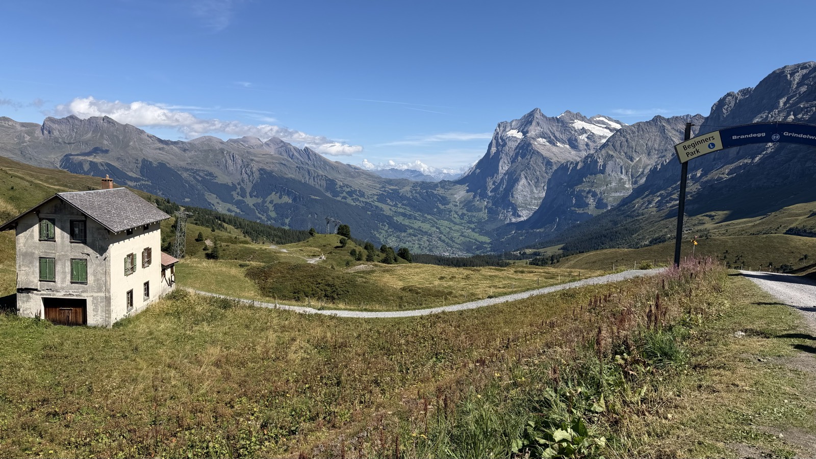 A stone house and alpine meadow with a gravel path, a yellow signpost and a snow-topped rock massif across the valley