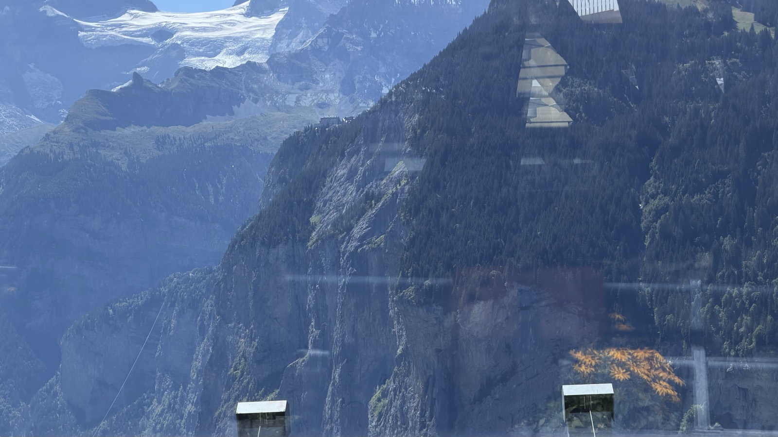 A sheer grey cliff wall across a deep valley with thin waterfalls falling down it, snow peaks behind, seen through a train window