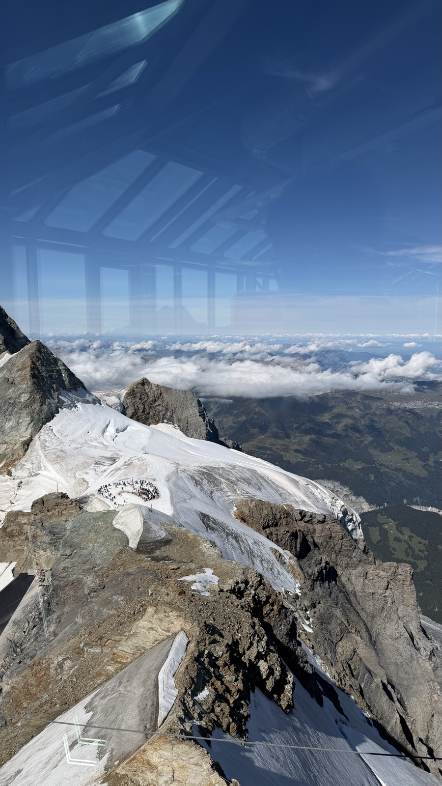 A glacier and rocky ridge seen from a high observation deck through glass, with a sea of cloud and distant lowlands beyond