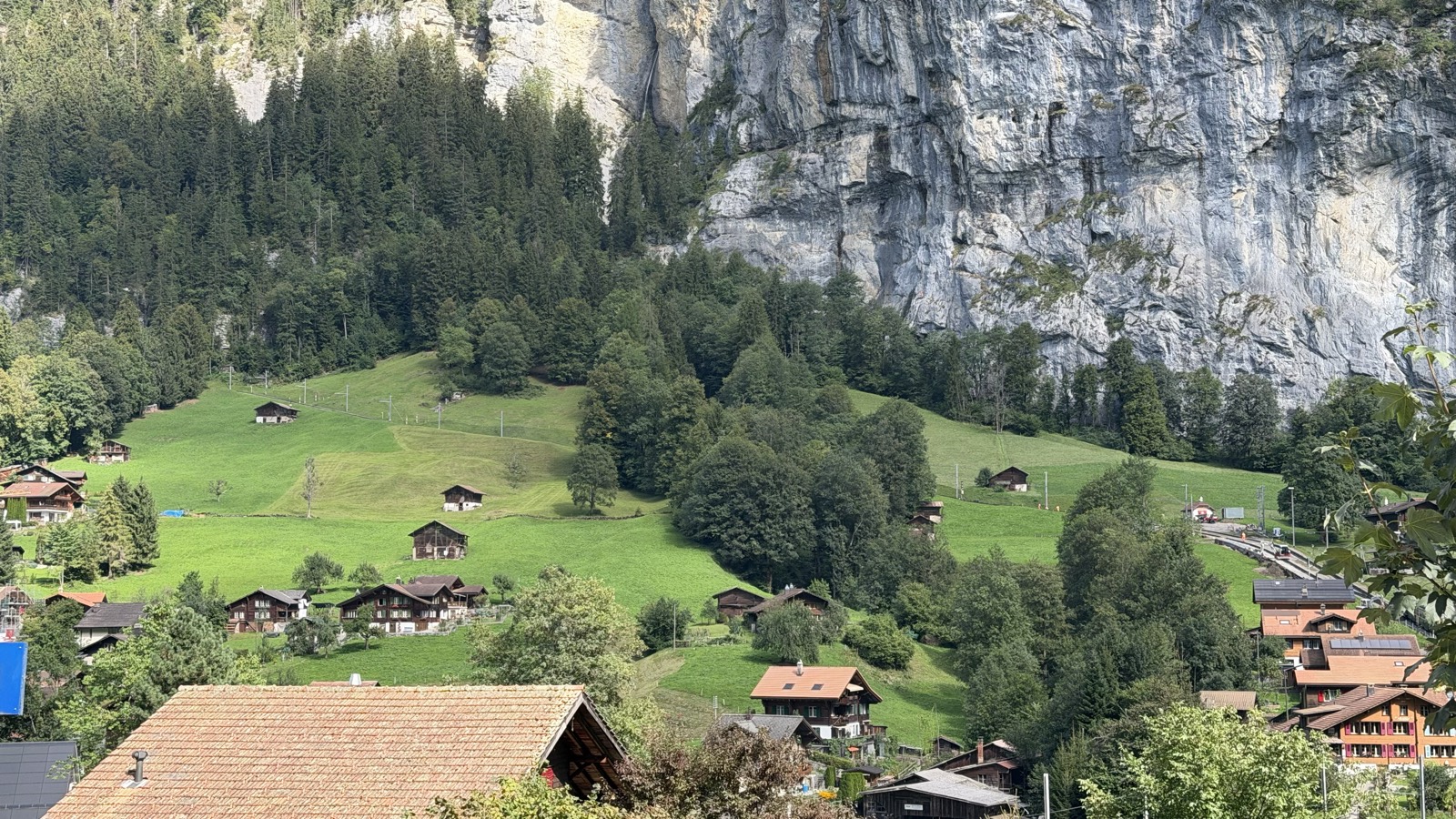 A Swiss village of chalets and green meadows below an enormous grey cliff wall, with a thin ribbon of waterfall running down the rock