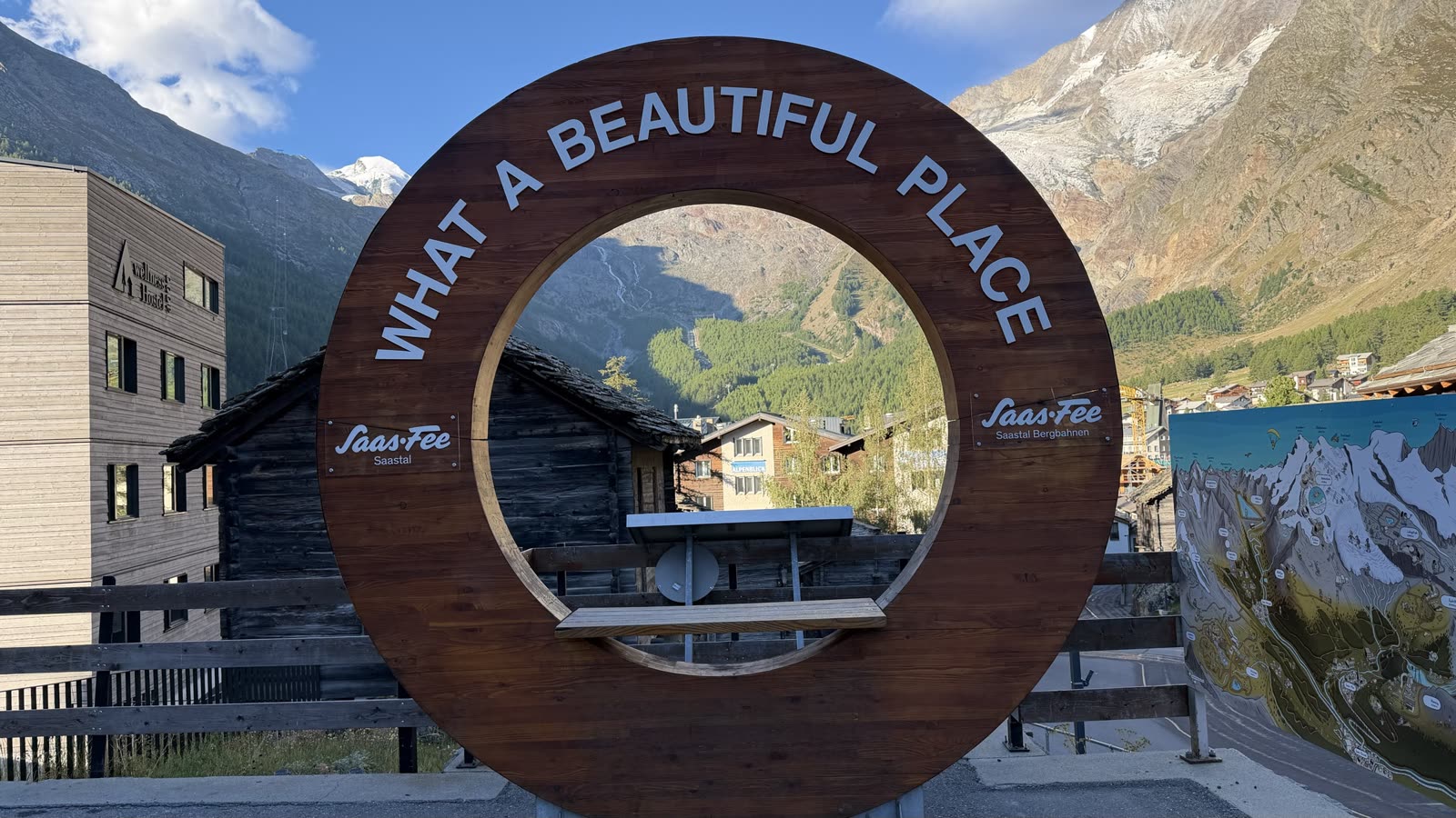 A wooden circular photo frame reading What a beautiful place, with Saas-Fee's chalets and a snow peak through it