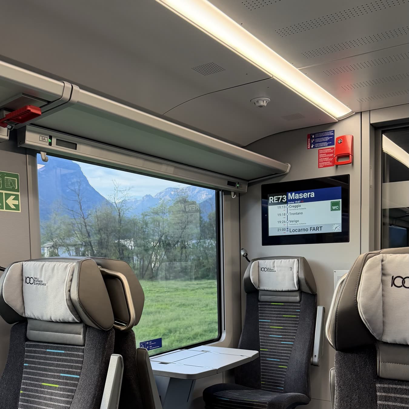 Empty seats and a small table inside a narrow-gauge train, a screen above showing the next stop