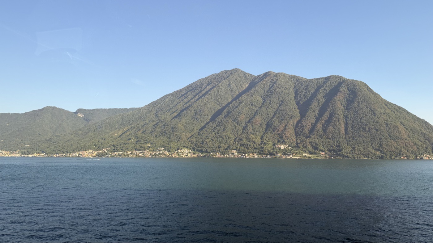 A wooded mountain falling to a line of lakeside villages across dark blue water under a clear evening sky
