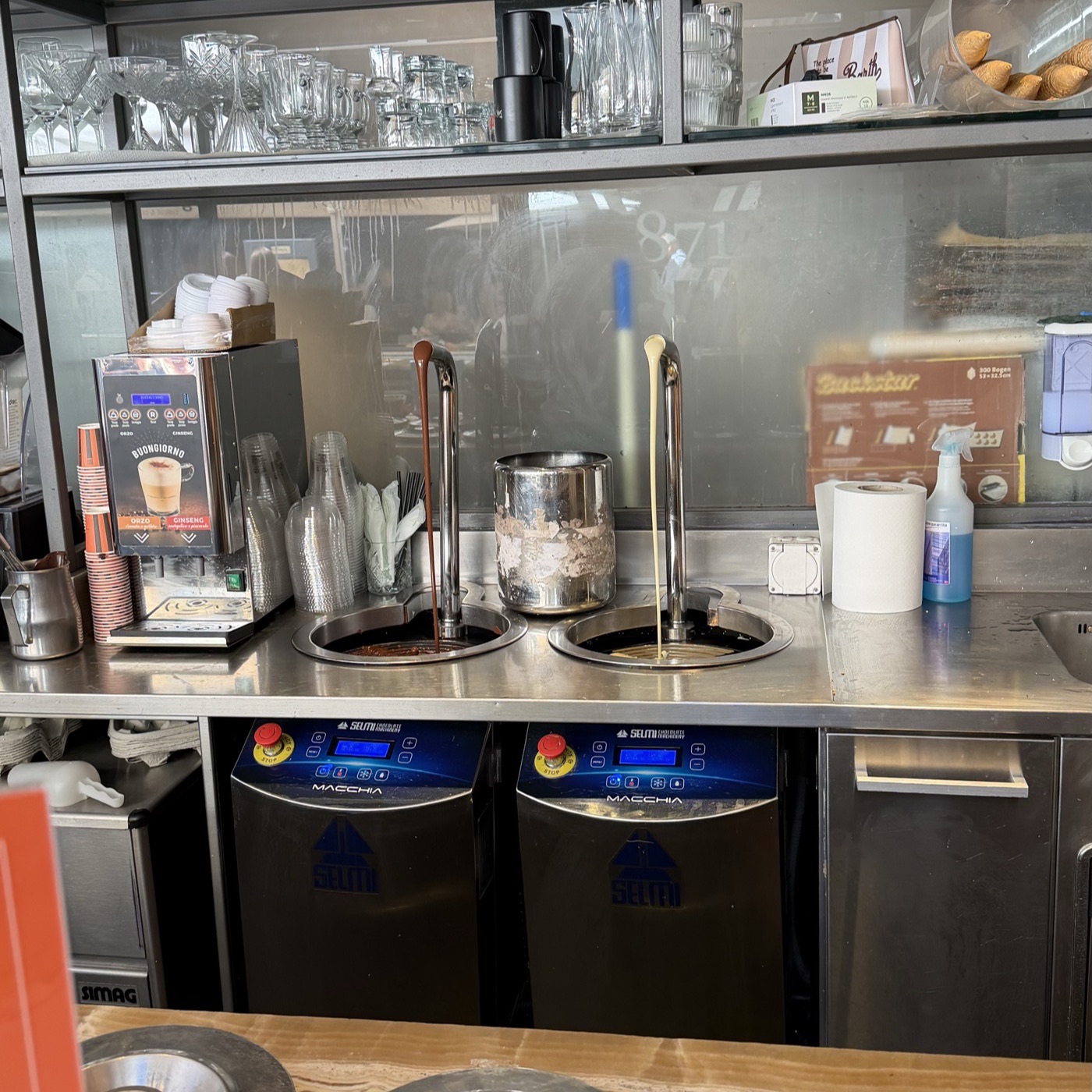 A gelato counter with steel lids over the tubs and two chocolate taps running behind it