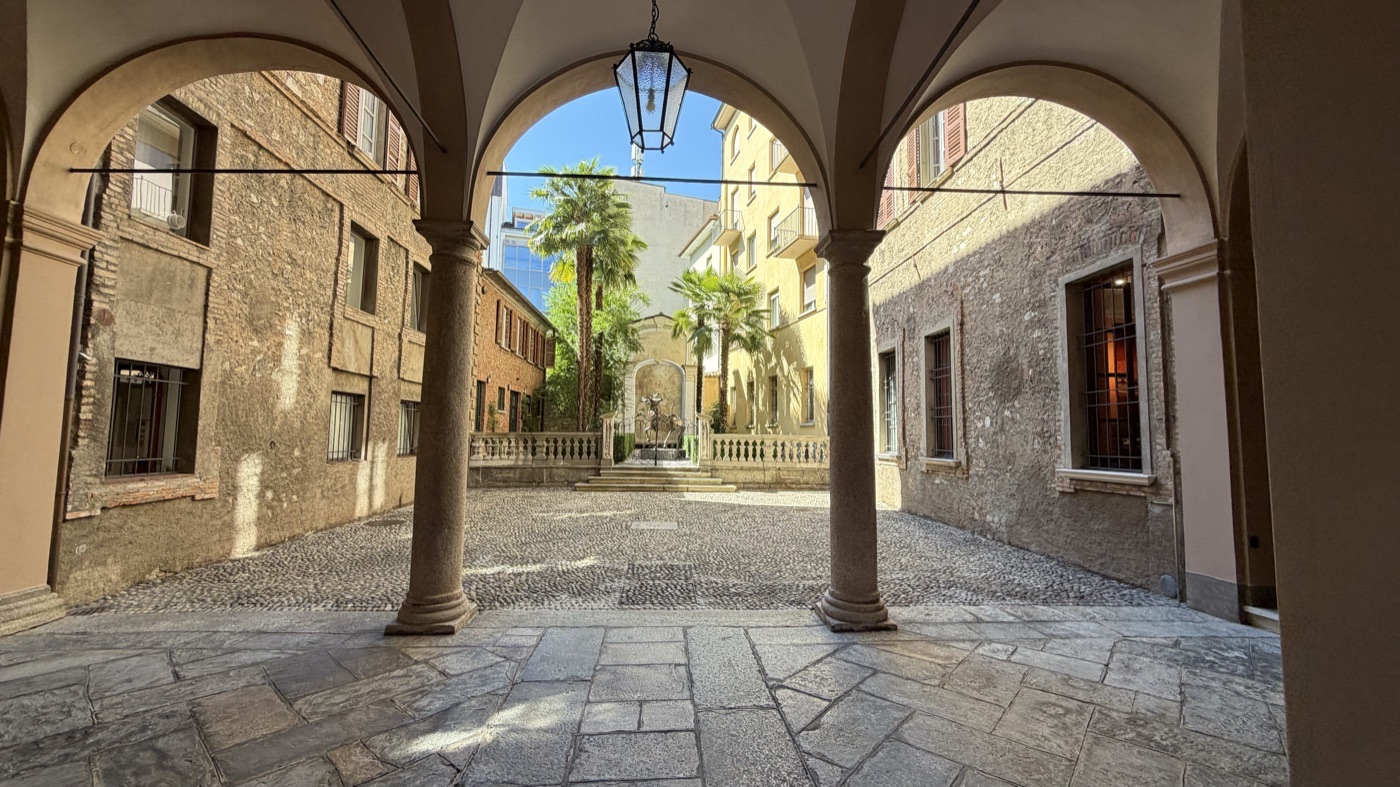 A vaulted arcade with a hanging lantern opening onto a courtyard with palm trees