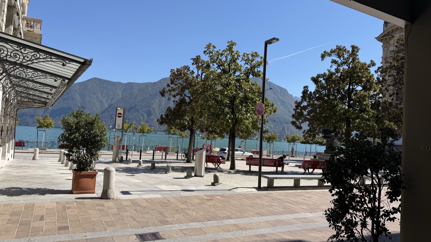 A lakeside promenade under plane trees with a mountain across the water