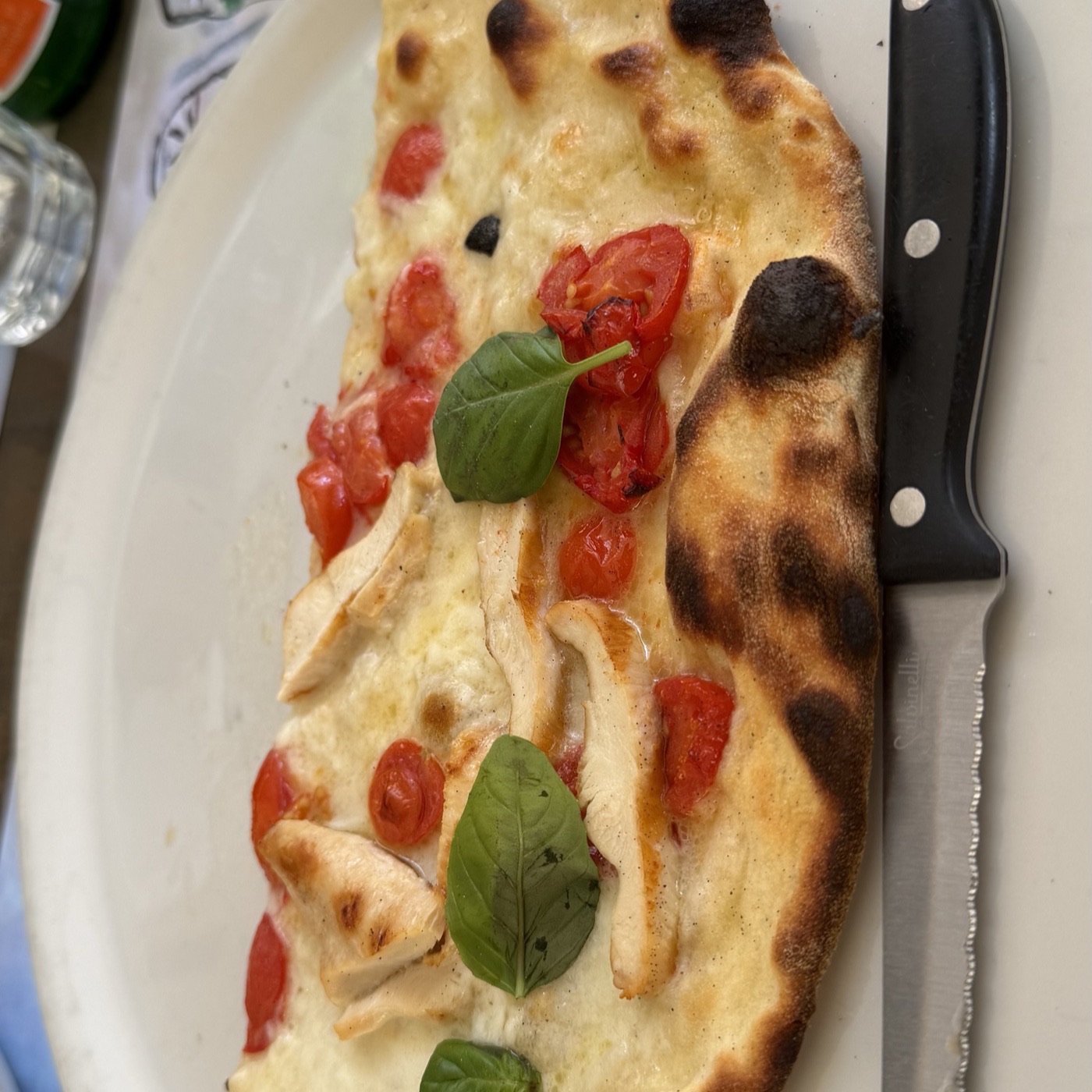 A white pizza with cherry tomatoes, chicken and basil on a white plate beside a steak knife