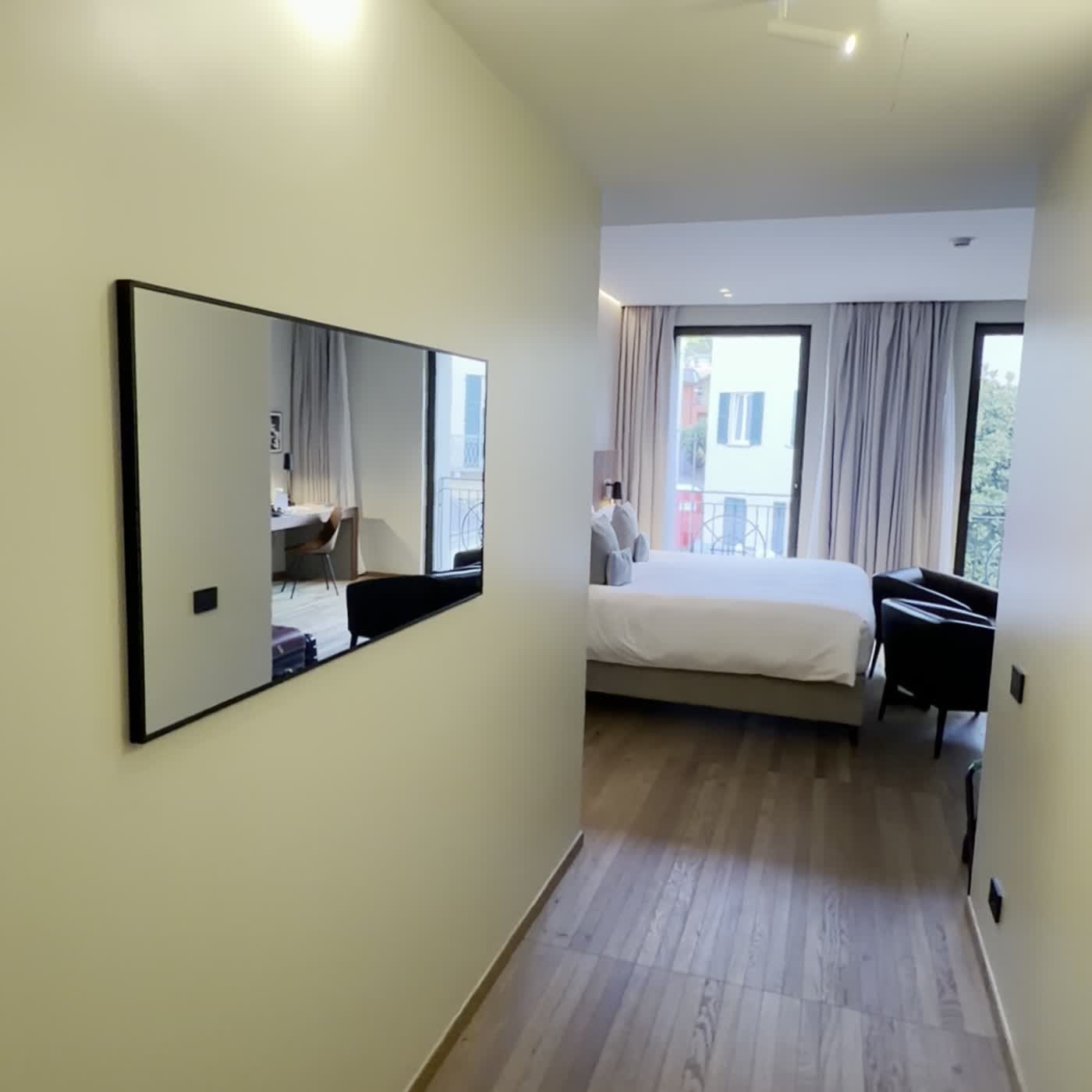 A hotel room seen from its entrance corridor, a white bed, two dark armchairs and tall windows onto trees and a balcony