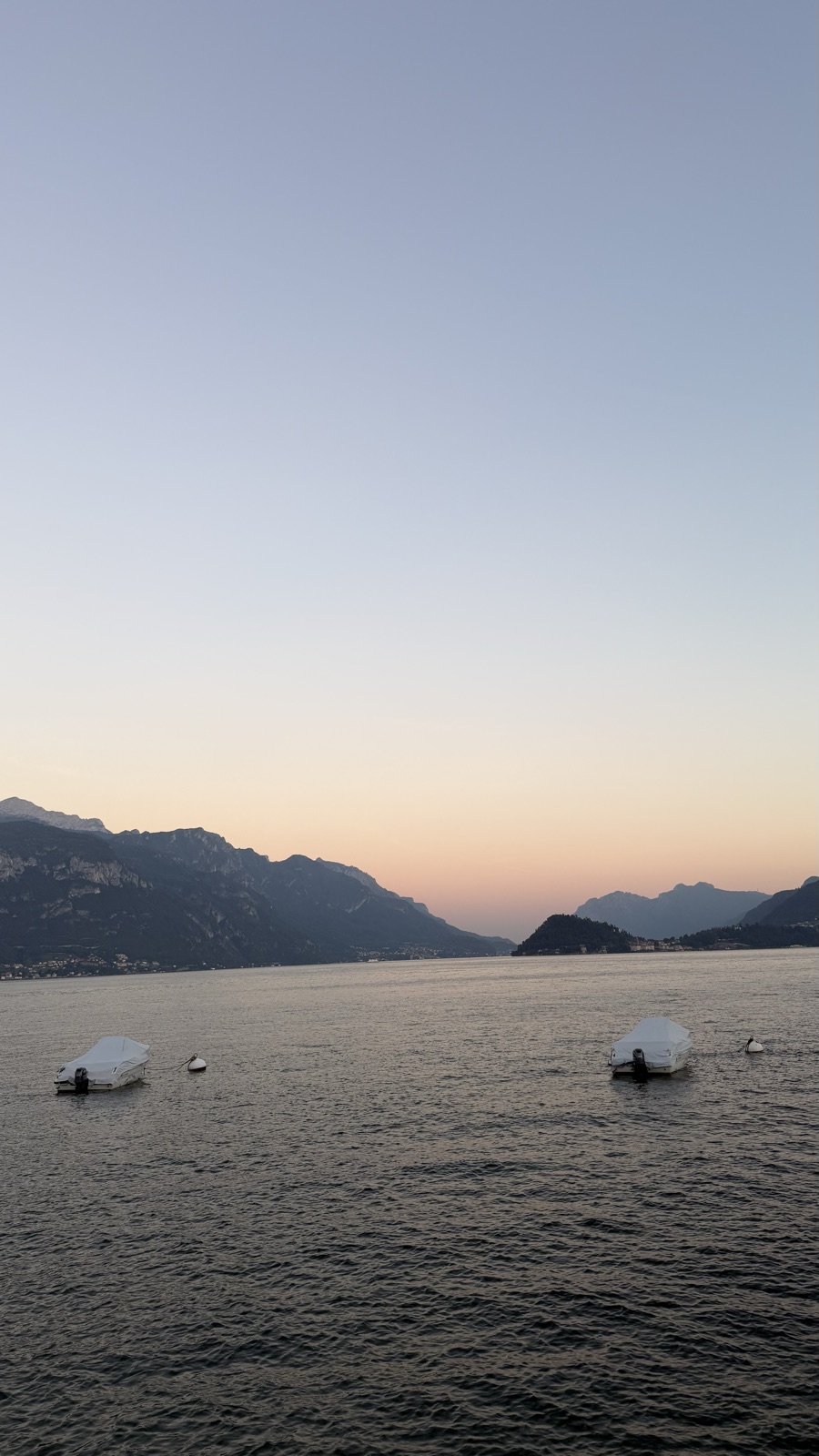 Small boats moored on a calm lake at dusk with mountains fading into the haze