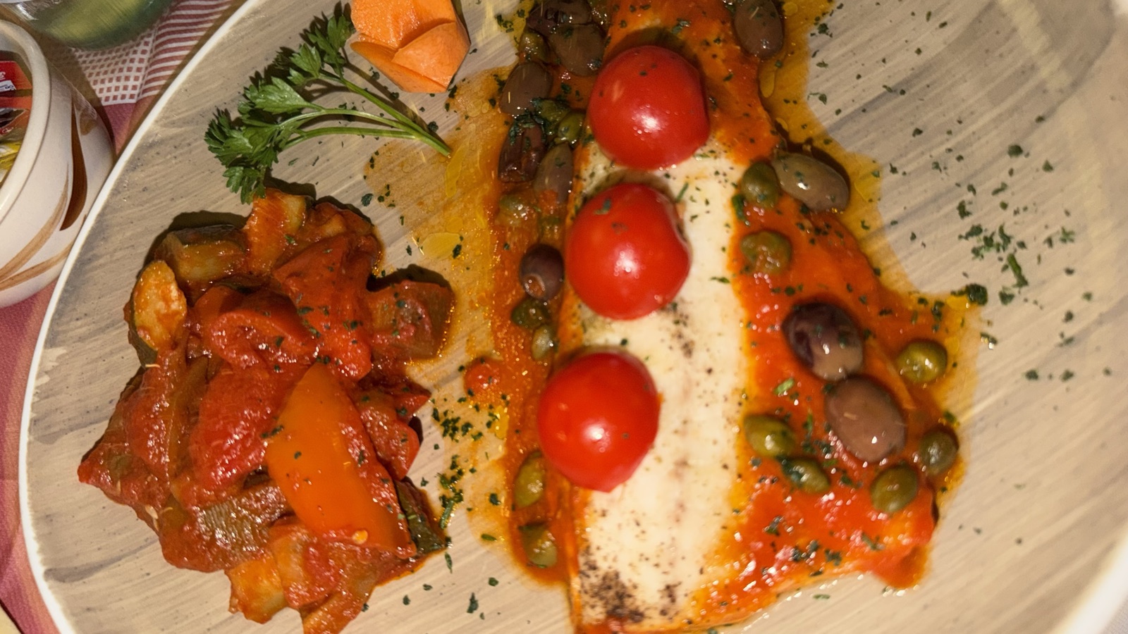 A plate of white fish with roasted peppers, cherry tomatoes and capers on a red checked tablecloth