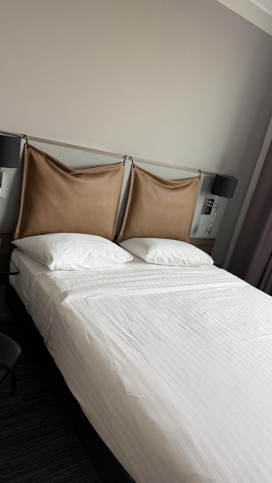 A hotel bed with white linen and tan leather headboard cushions beside a window