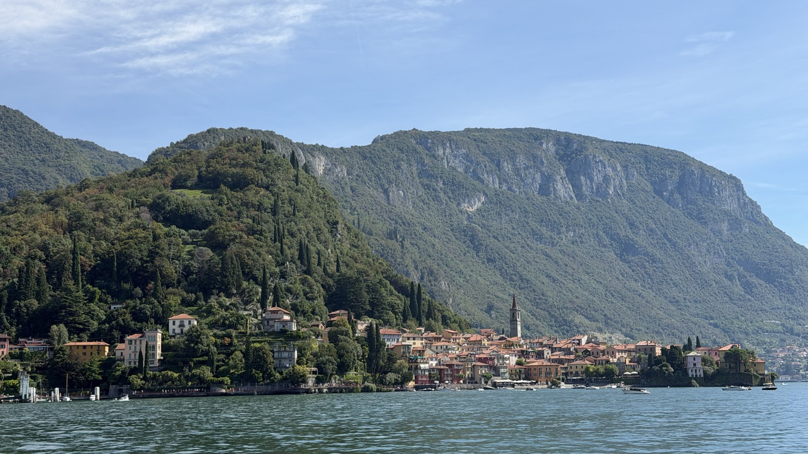A lakeside village of ochre houses climbing a wooded hill, seen from the water