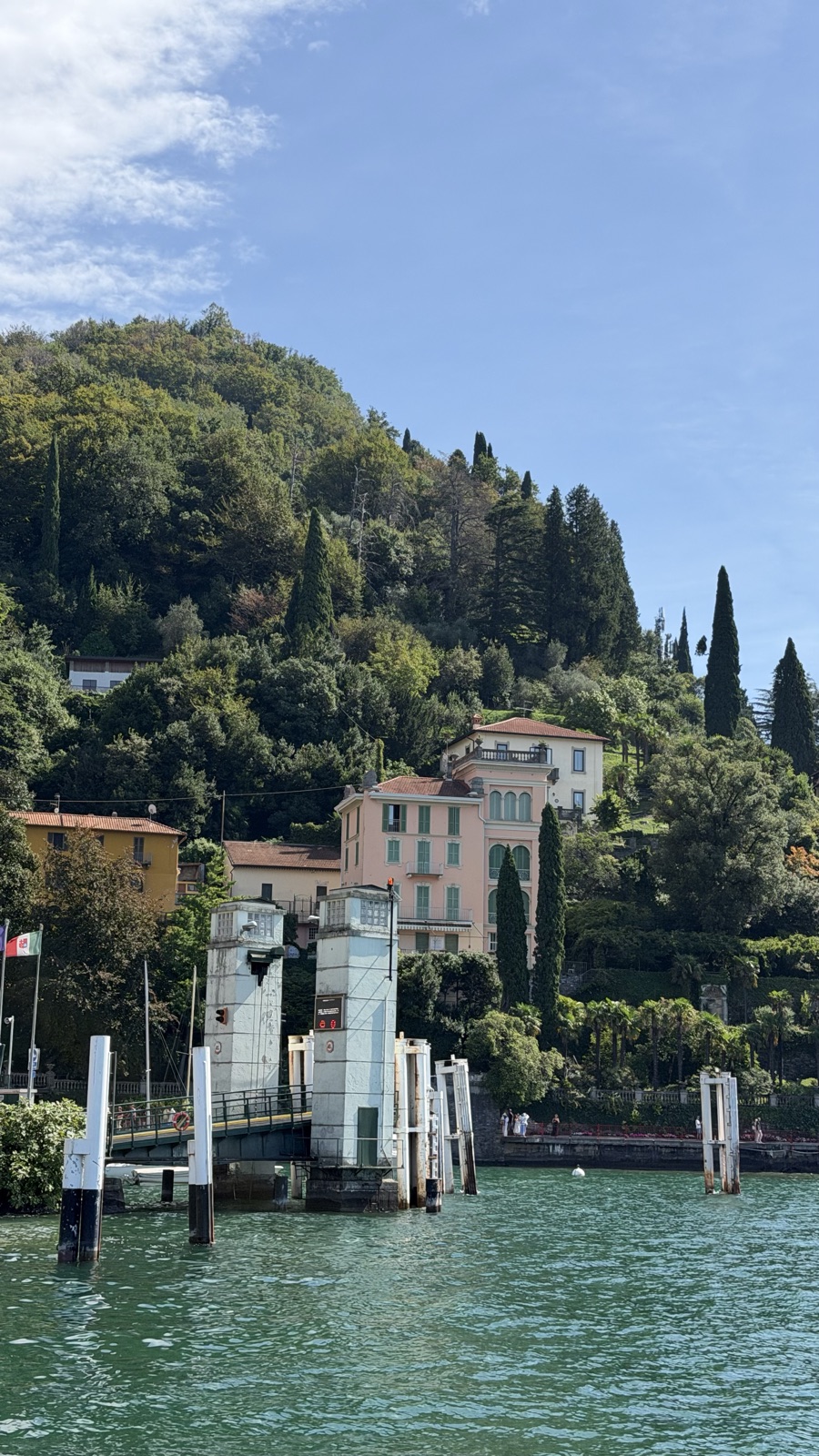 Mooring posts and a stone pier below a hillside of villas and cypress trees