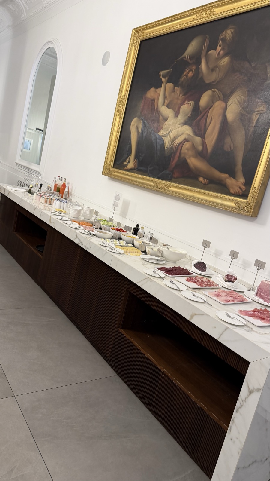 A breakfast buffet counter with pastries and cold cuts under a large gilt-framed painting