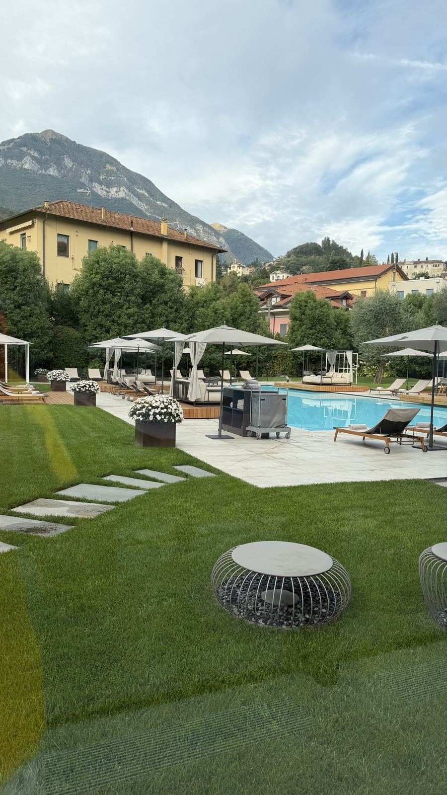 A hotel pool and lawn with sun loungers, a village and green hills beyond