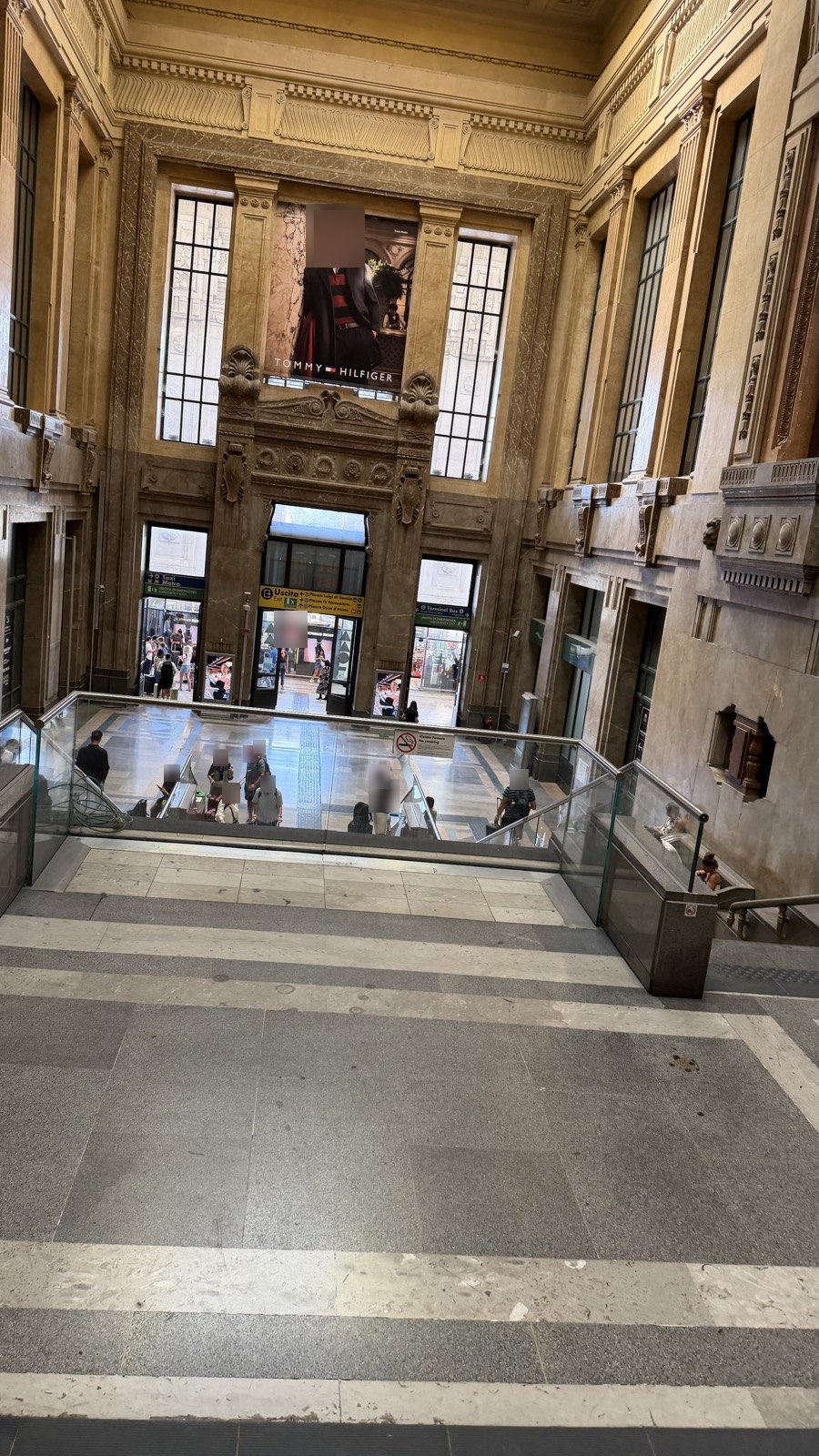 The grand stone entrance hall of Milano Centrale seen from the top of the stairs, with travellers crossing the floor below