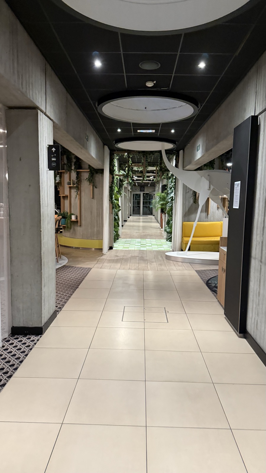 A long, bright hotel corridor with a tiled floor and a planted arch at the far end
