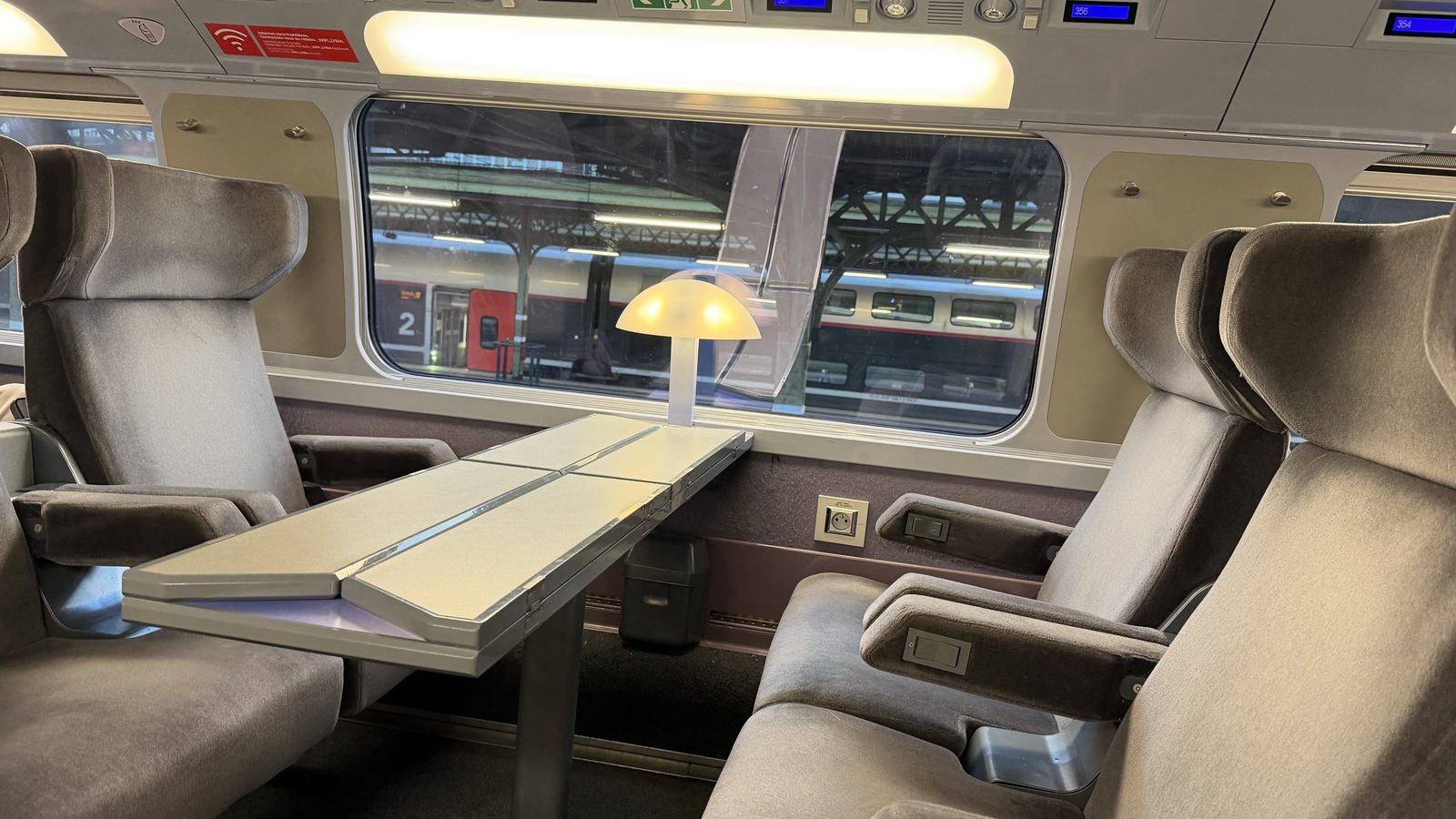 Two wide first-class seats on the TGV Lyria facing a small table, with the lamp on and the platform outside the window