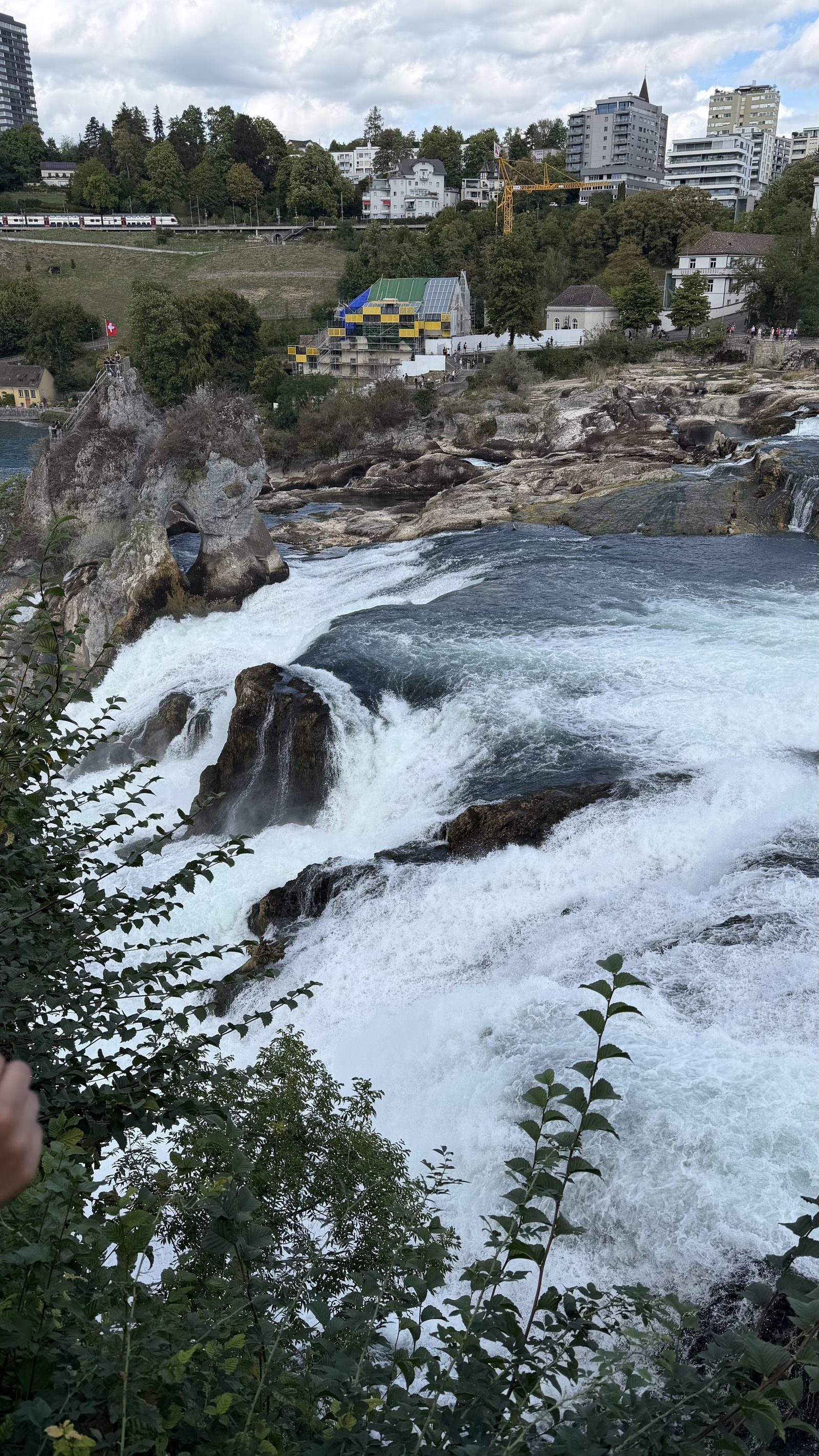 The Rhine Falls from the walkways, white water pouring over the rocks with the town and a castle on the hill above