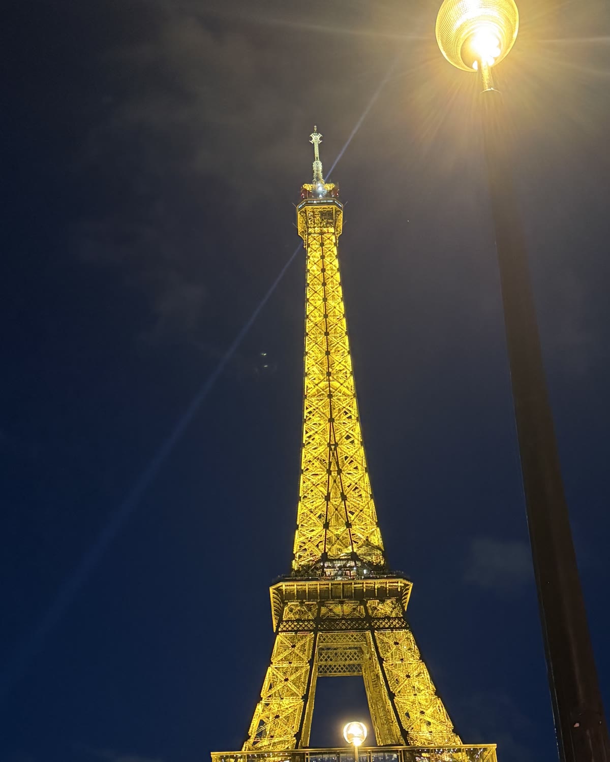 The Eiffel Tower lit at night, its beacon sweeping the sky, photographed from the foot of the tower