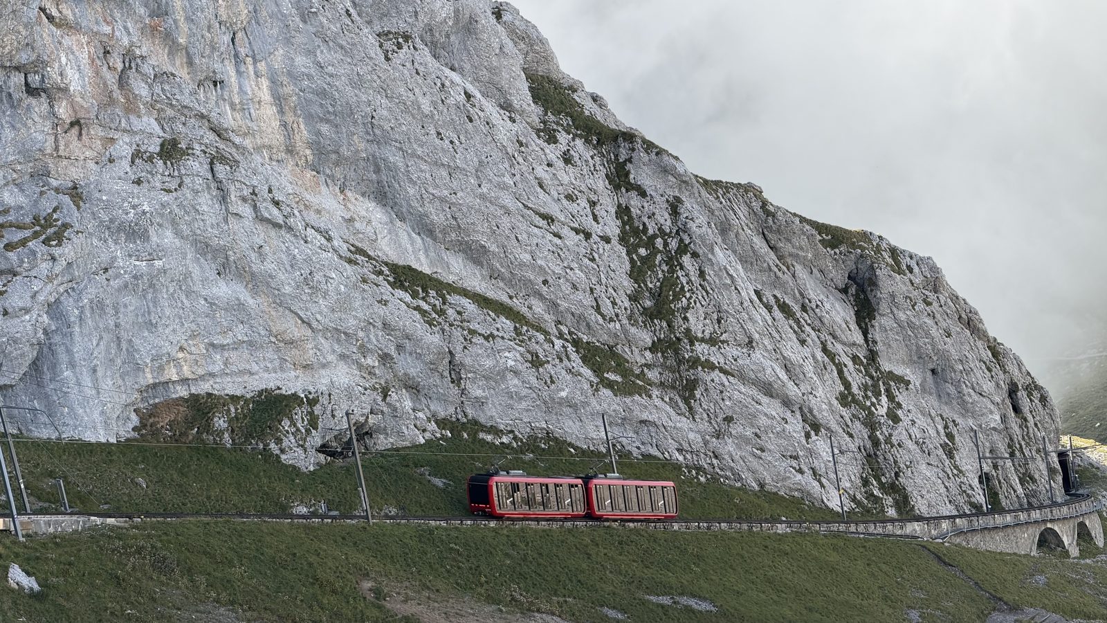 A red two-carriage cogwheel train on a narrow track cut into a grey limestone face on Pilatus