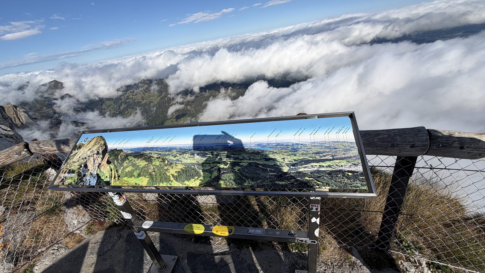 A metal panorama board at the Pilatus summit naming the peaks, with cloud filling the valley behind it